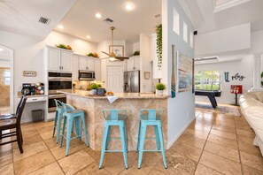 A modern kitchen with white cabinets, a central island with bar stools, and a pool table in the adjacent living area.