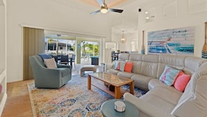 Ein geräumiges Wohnzimmer mit grauem Sofa, blauem Sessel und Couchtisch. Eine Deckenventilator, ein großes Fenster und ein Gemälde an der Wand sind vorhanden.