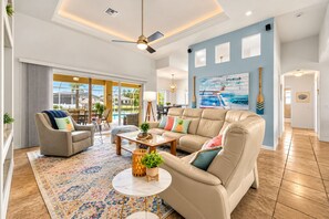A spacious living room with a large beige sofa, a coffee table, and a patterned rug. There is a painting on the wall and a view of the outdoors through large windows.