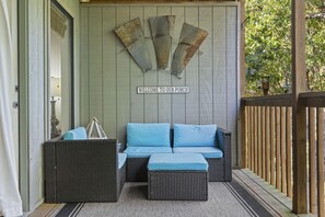 A porch with a wicker sofa and cushions, a wooden wall with a 'WELCOME TO OUR PORCH' sign, and a wooden railing.