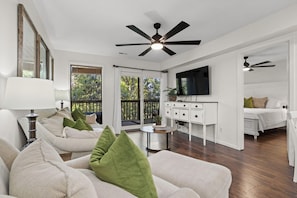 A spacious living room with a ceiling fan, a television, and a balcony with a view of trees.
