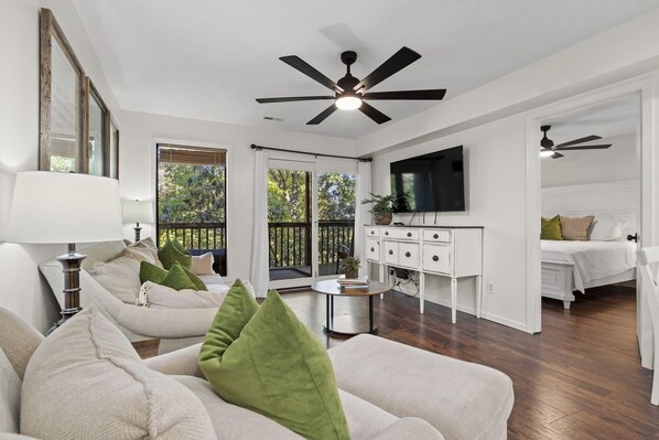 A spacious living room with a ceiling fan, a television, and a balcony with a view of trees.