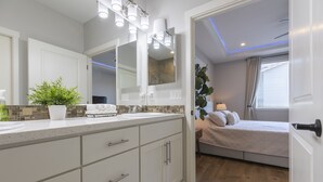A modern bathroom with a large mirror, multiple lights, a potted plant, and a view into a bedroom with a bed and curtains. - Cozy new built home 10 minutes away from Portland (Vancouver)