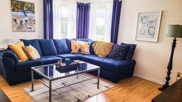 Un salón con un sofá seccional azul, una mesa de centro de vidrio y una alfombra. Hay dos ventanas con cortinas moradas, un cuadro en la pared y una lámpara sobre una mesita.