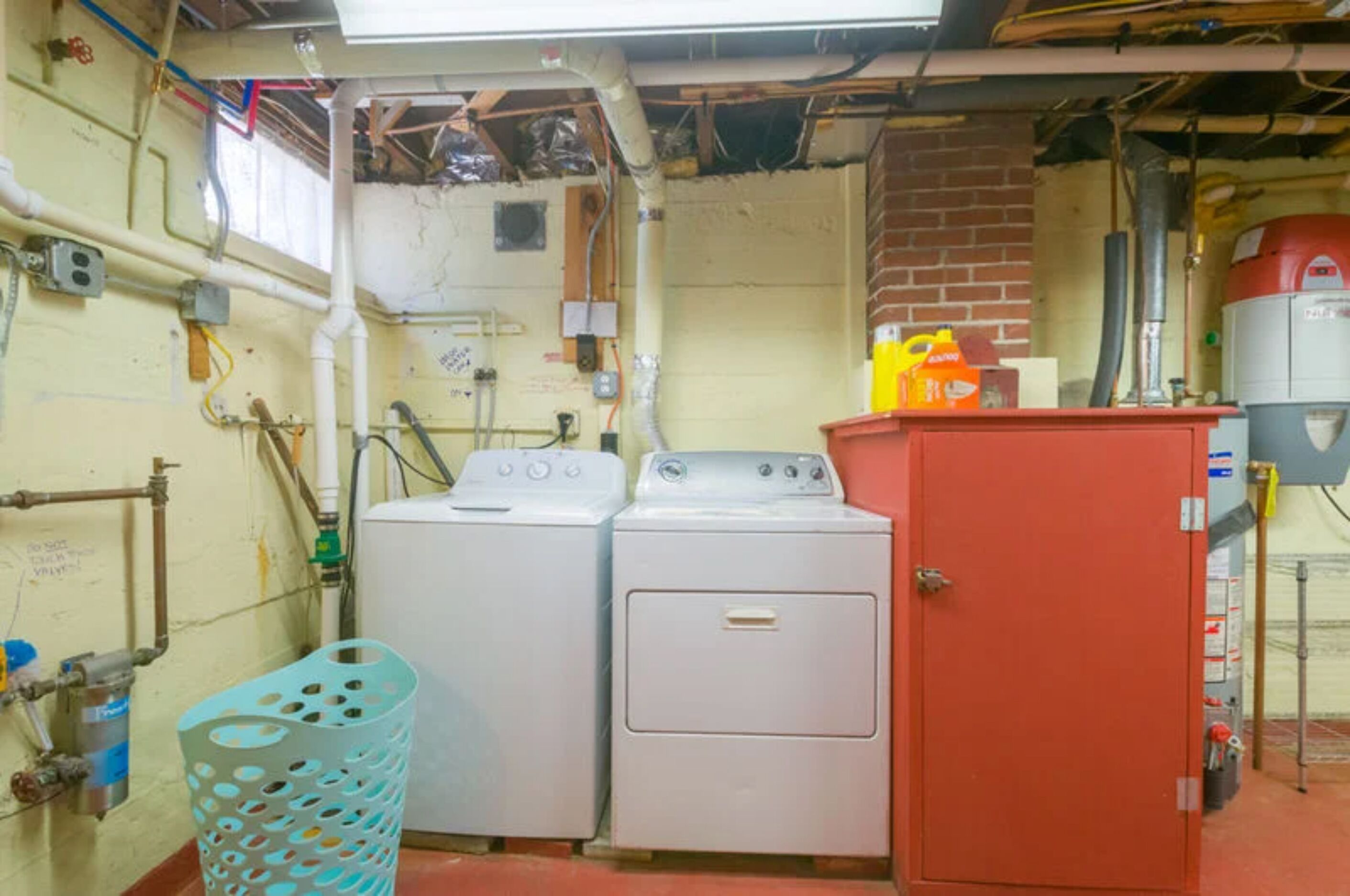 A laundry room with two washing machines, a red utility cabinet, and various pipes and electrical outlets on the walls.