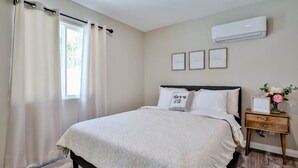 A bedroom with a bed, a nightstand, a window with curtains, and an air conditioning unit on the wall. - Private Pool & Hot Tub - Minutes from Disneyland - Comfort & Convenience (Anaheim)