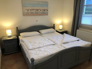 A bed with two pillows and a quilt, flanked by nightstands with lamps, in a room with a painting on the wall and a window with blinds.