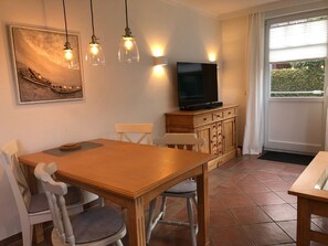 A dining area with a wooden table and chairs, a television on a wooden cabinet, and pendant lights above the table.