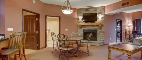 A living room with a stone fireplace, a dining table with chairs, and a television mounted on the wall.
