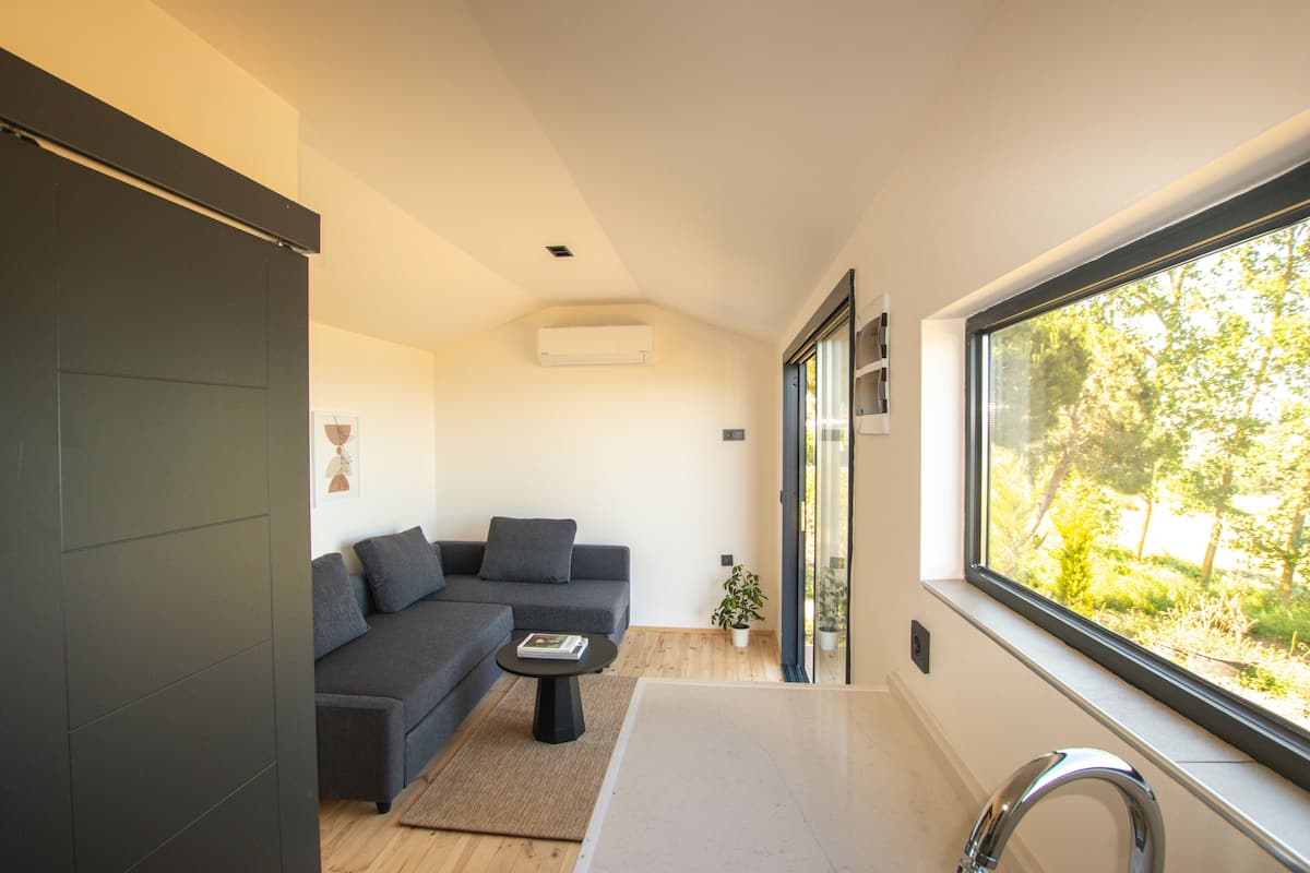 A modern living room with a grey sofa, a small round table, and a large window offering a view of greenery.