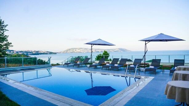 Zona de piscina con sillones de descanso y sombrillas, con vistas a un paisaje urbano y al mar.