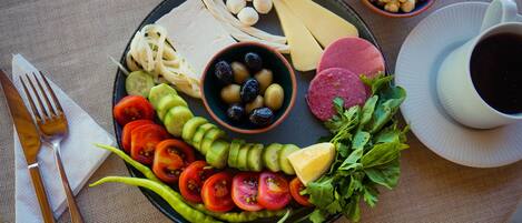 A plate with sliced tomatoes, cucumbers, cheese, olives, and meat, accompanied by a cup of coffee and a bowl of chickpeas.