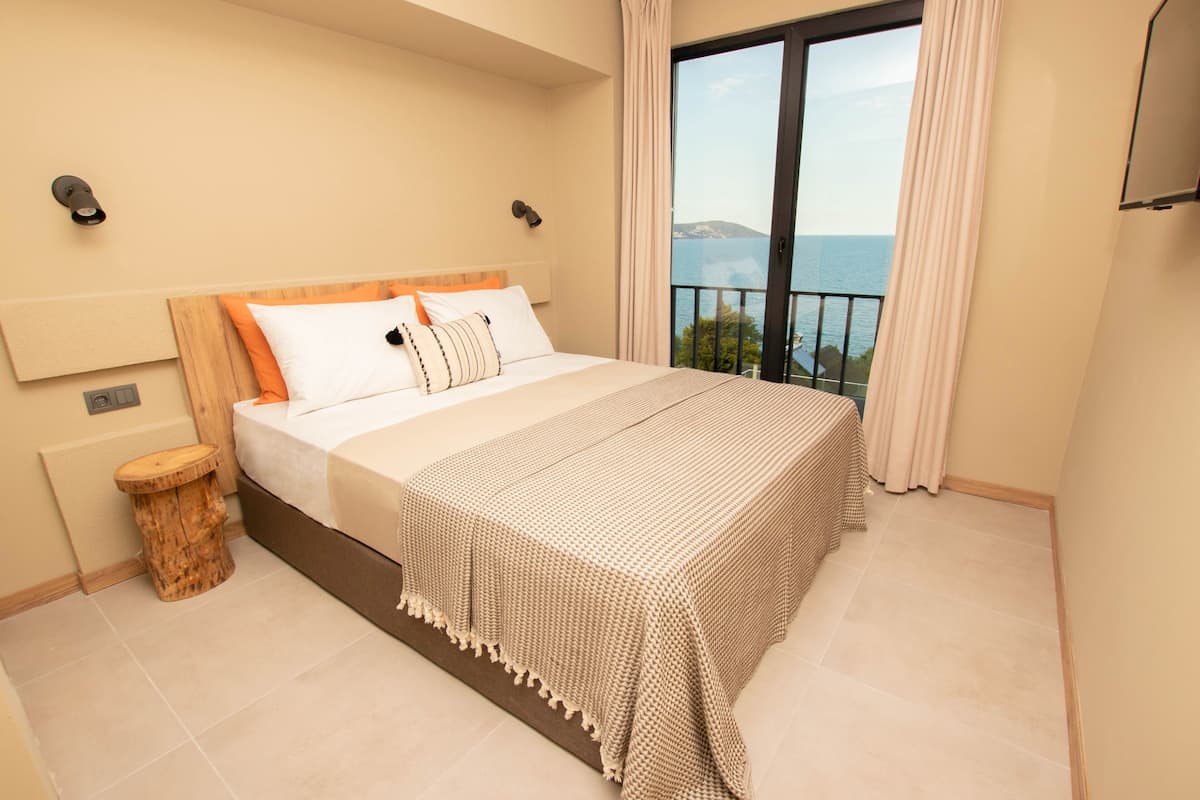 A bedroom with a large bed, a wooden nightstand, a view of the sea through a balcony, and a ceiling-mounted light fixture.