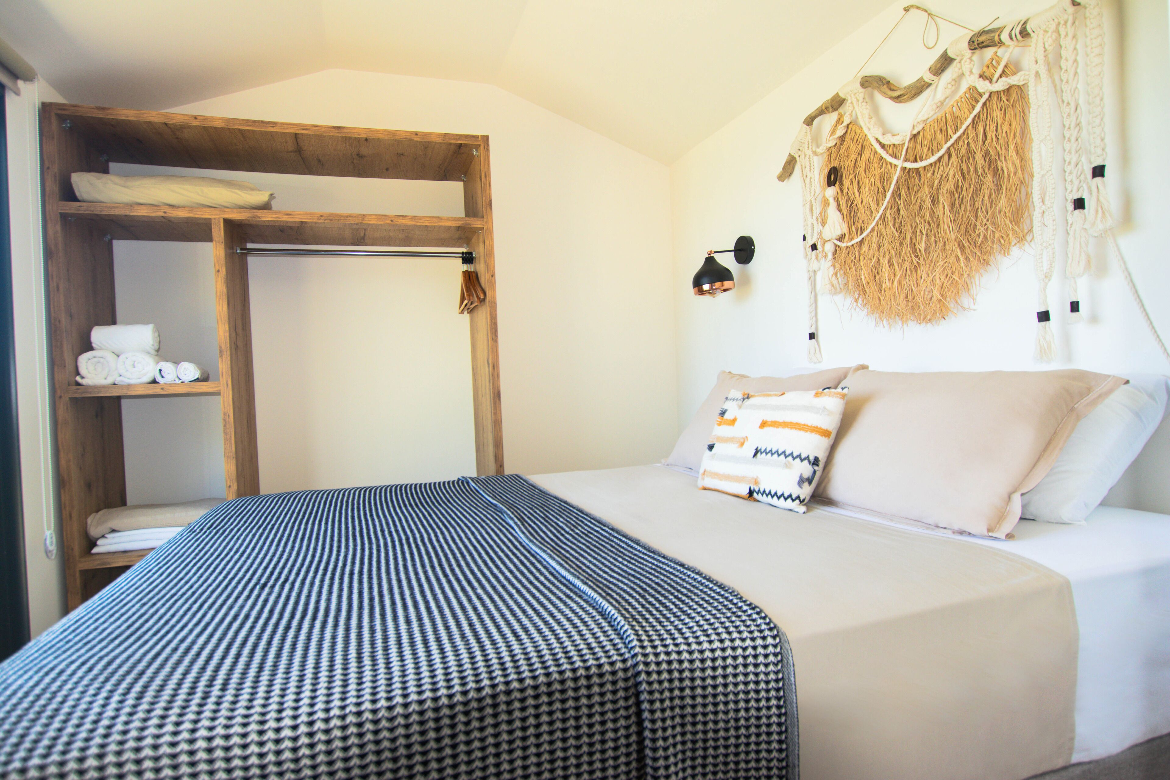 A room with a bunk bed, a wooden shelf with towels, a wall-mounted lamp, and a macrame wall hanging.