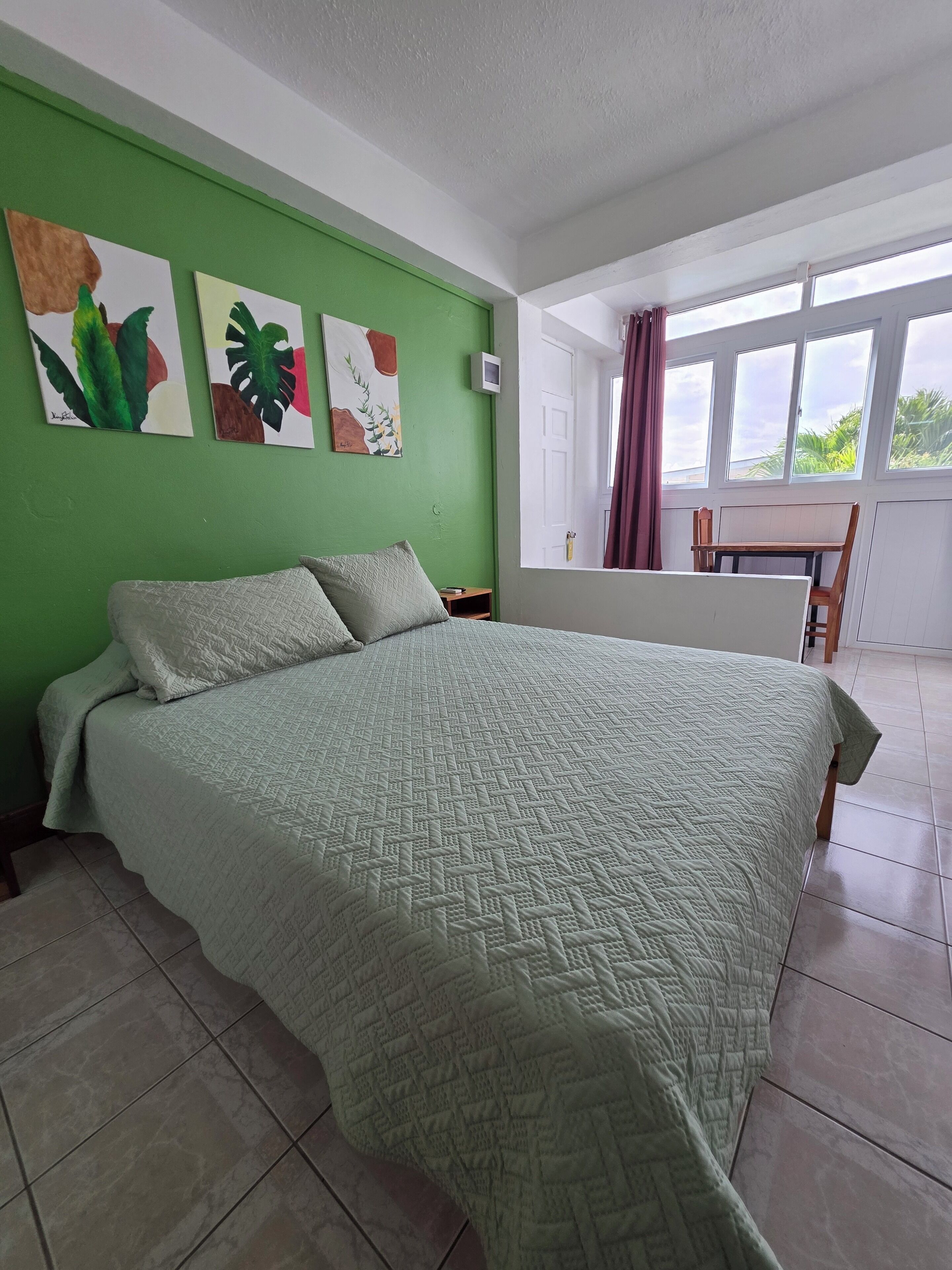 Une chambre avec un grand lit, des murs verts et des œuvres d’art. Il y a une fenêtre avec des rideaux et une vue sur la verdure à l’extérieur.