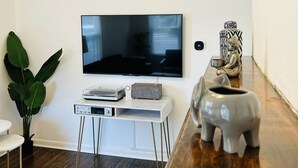 A living room with a flat-screen TV mounted on the wall, a white entertainment unit with a turntable and speakers, a wooden coffee table with decorative items, and a large green plant.