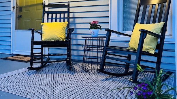A porch with two rocking chairs and a small table, a potted plant, and a door with a number above it.