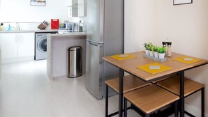 Ett modernt kök med ett kylskåp i rostfritt stål, en tvättmaskin och en sopbehållare. Det finns ett träbord med gula bordsunderlägg och en inramad tavla på väggen.