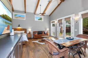 A spacious living area with a wooden ceiling, a sofa, a dining table, wicker chairs, and a large window with a view of a lake.