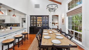 Una cocina amplia con una isla grande, una mesa de comedor de madera dispuesta para seis personas y vistas al exterior a través de grandes ventanales.