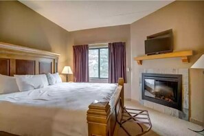 A hotel room with a large bed, a television on a shelf, a fireplace, and a window with curtains.
