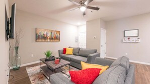 A living room with a grey sofa, a coffee table, a TV, a ceiling fan, and a painting on the wall. - Luxury Furnished Rental in Conway, AR Unit 206 (Conway)