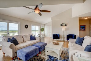 A spacious living room with a beige sofa, blue ottoman, and white coffee table. There is a ceiling fan, a clock on the wall, and a view of the ocean through large windows.