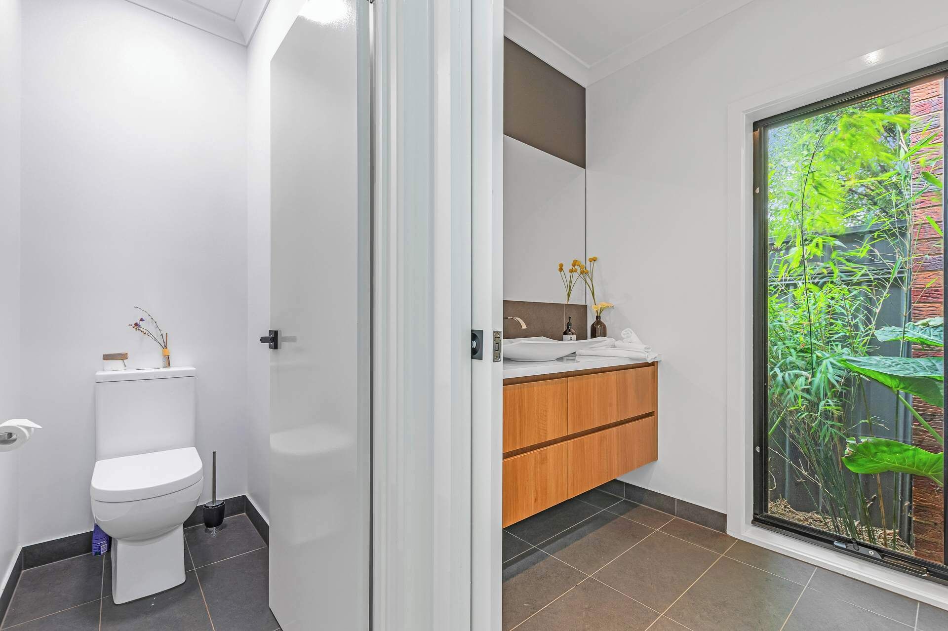 A modern bathroom with a toilet, a vanity with a mirror, and a view of greenery through a glass door.