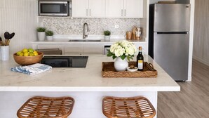 A modern kitchen with white cabinets, a stainless steel refrigerator, a microwave, and a central island with stools. - Island Life Retreat-Ocean Front-Beach Access (Hauula)