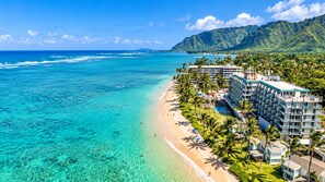 A beachfront hotel with multiple buildings, a pool, and a clear view of the ocean and mountains.