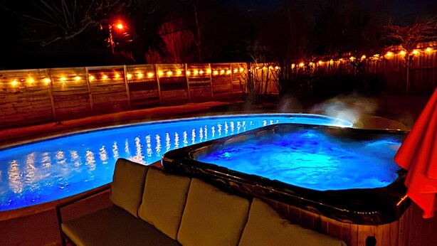 Ein Whirlpool mit Holzkante und integriertem Sitzplatz, umgeben von einer Holzterrasse mit Lichterketten.