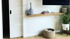A modern living room with a flat-screen TV mounted on the wall, a white door, a wooden shelf with decorative items, and a wicker basket on the floor.