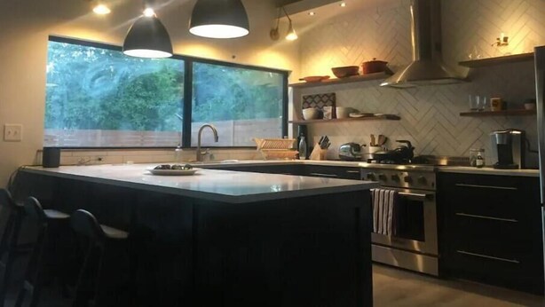 A modern kitchen with a large island, pendant lighting, a view of greenery outside, and a herringbone-patterned backsplash.