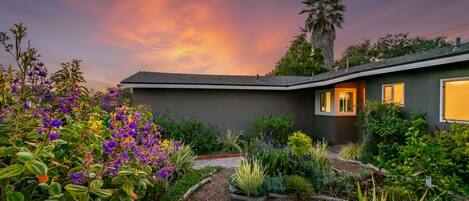 A modern house with a well-maintained garden, a palm tree, and a sunset sky.