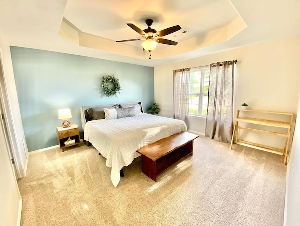 A bedroom with a bed, a nightstand, a bench, a ceiling fan, a window with curtains, and a wall-mounted wreath. - Shred Shed- Sleeps 10- Near Trails- Bentonville (Bentonville)