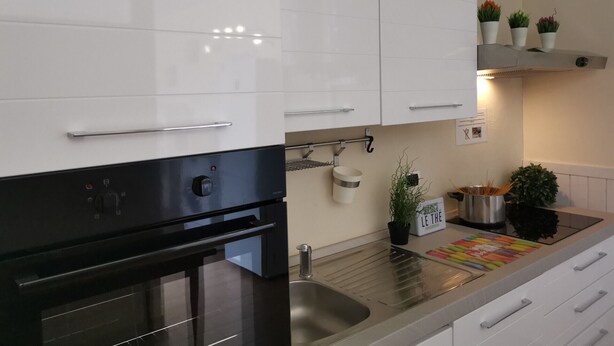 A modern kitchen with white cabinets, a stainless steel sink, and a black oven. There are potted plants on the countertop and a small sign that reads "LE THE".