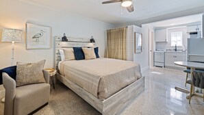 A bedroom with a bed, a chair, a nightstand, a ceiling fan, and a view into the adjacent kitchen.