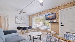 Una sala de estar moderna con un sofá gris, una mesa redonda de centro y un sillón azul estampado. Hay un televisor en una pared de madera, un ventilador de techo y una ventana con persianas.