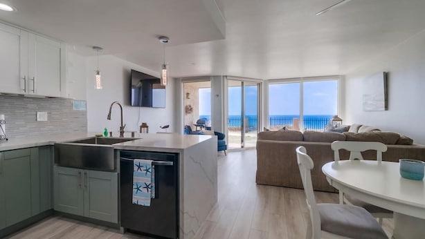 Una cocina moderna con una isla central, un comedor con una mesa y sillas blancas, y una sala de estar con sofá y vistas al mar.