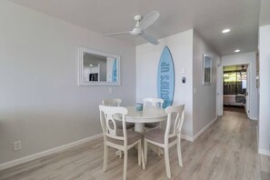 A dining area with a round table and four chairs, a ceiling fan, a surfboard decoration, and a mirror on the wall. - Private Beach Access | Oceanfront | Pool & Spa (Solana Beach)