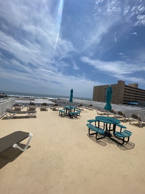 A rooftop terrace with sun loungers, picnic tables, and umbrellas, overlooking the ocean and a building in the background.