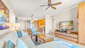 A modern living room with a sofa, coffee table, and TV. There is a dining area with chairs and a table, and a kitchen in the background.