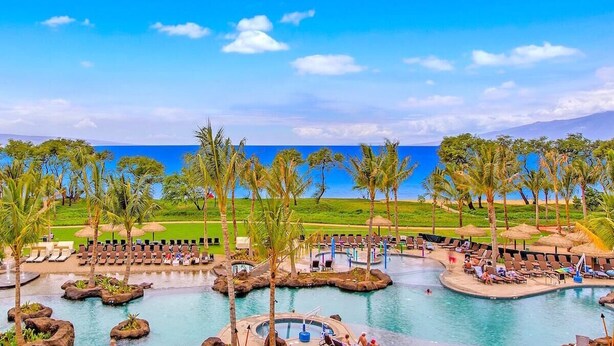 A resort pool area with lounge chairs and palm trees, overlooking a beach and ocean.