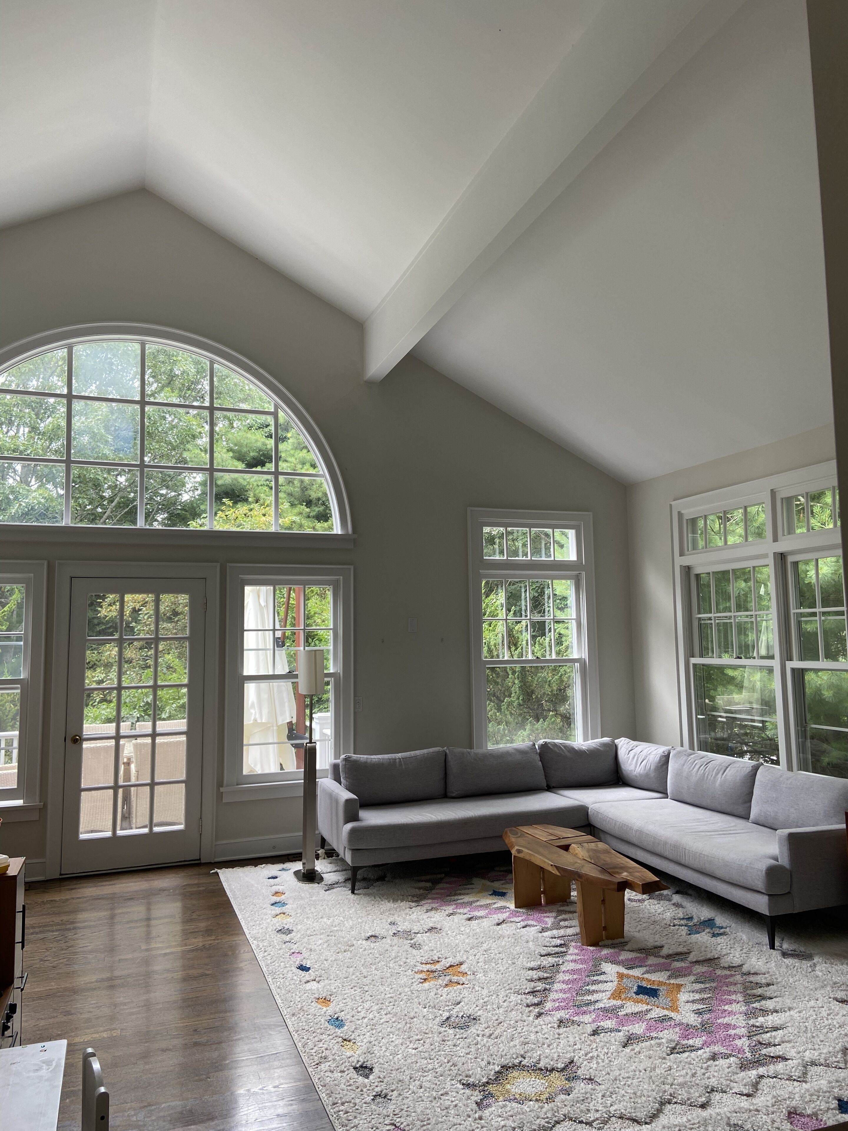 A spacious living room with a high ceiling, large windows, a grey sectional sofa, a wooden coffee table, and a patterned rug.