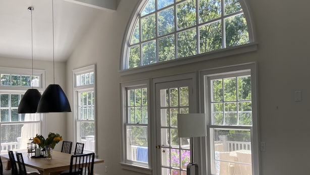 A spacious room with a high ceiling, large windows, a dining area with a table and chairs, and a gray sofa. - Pristine Southampton Home (Water Mill)