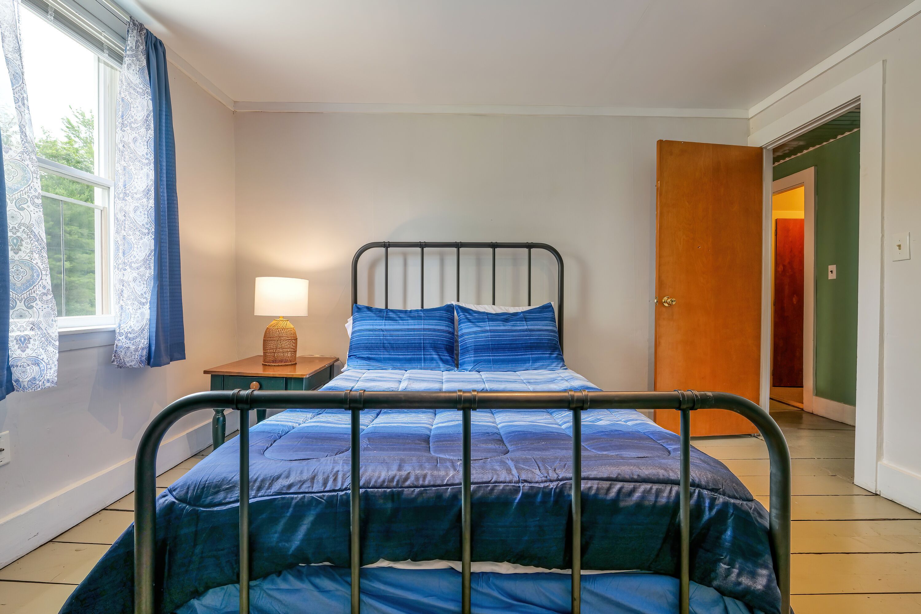 Une chambre à coucher avec un lit en métal, une couvre-lit bleue, une lampe de chevet et une fenêtre avec des rideaux.