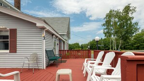 A covered outdoor deck with white plastic chairs and a small table, a red railing, and a house with white siding and brown shutters.