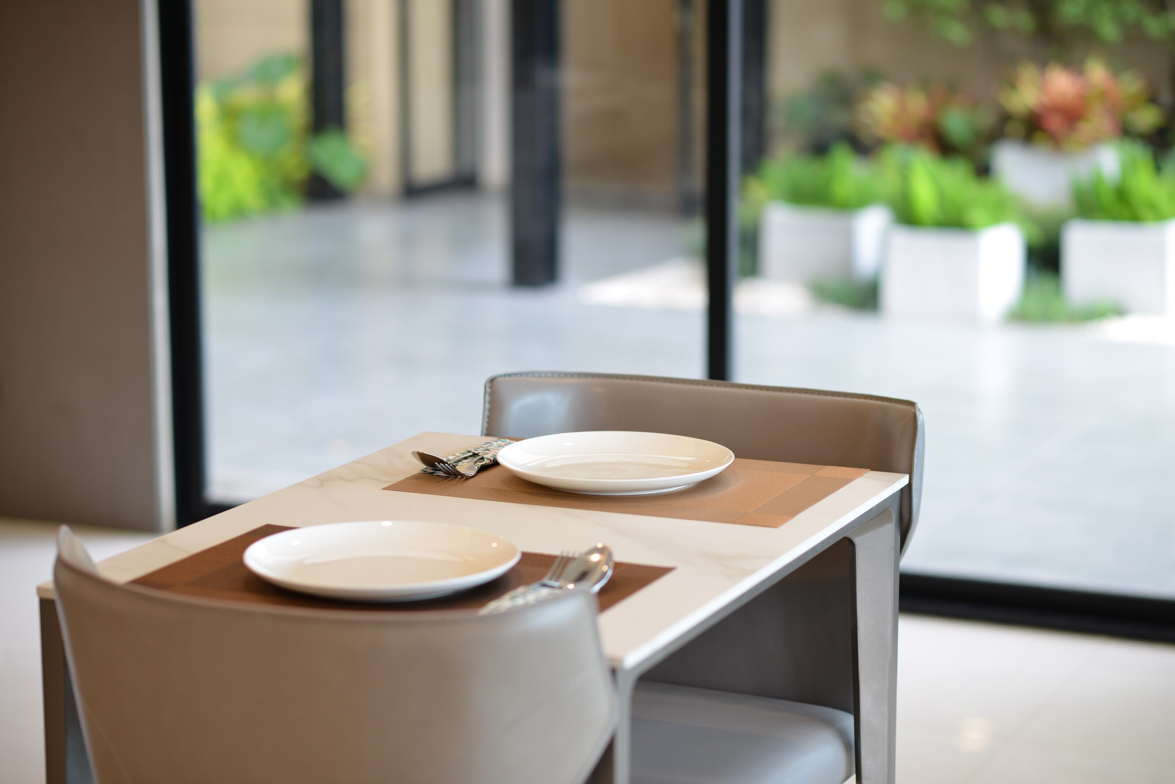 A dining table with two plates, cutlery, and a napkin, set in a modern interior with a view of greenery outside.