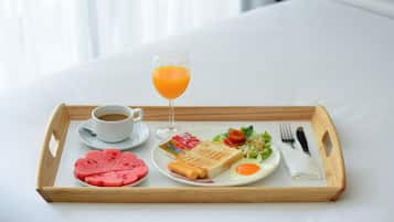 Ein Frühstücksbrett mit einer Kaffeetasse, einem Orangensaftglas, einem Wassermelonennstück, einem Sandwich, einem Spiegelei und einem kleinen Salat auf einem Bett.