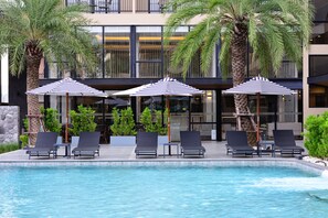 A pool area with lounge chairs and umbrellas, surrounded by palm trees and modern buildings. - Grand BS Airport Hotel Suvarnabhumi (Bangkok)